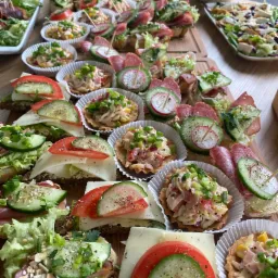 Różnorodne kanapki i tartaletki na drewnianej desce, udekorowane świeżymi warzywami, wędlinami i ziołami, tworzące apetyczną kompozycję przekąsek.