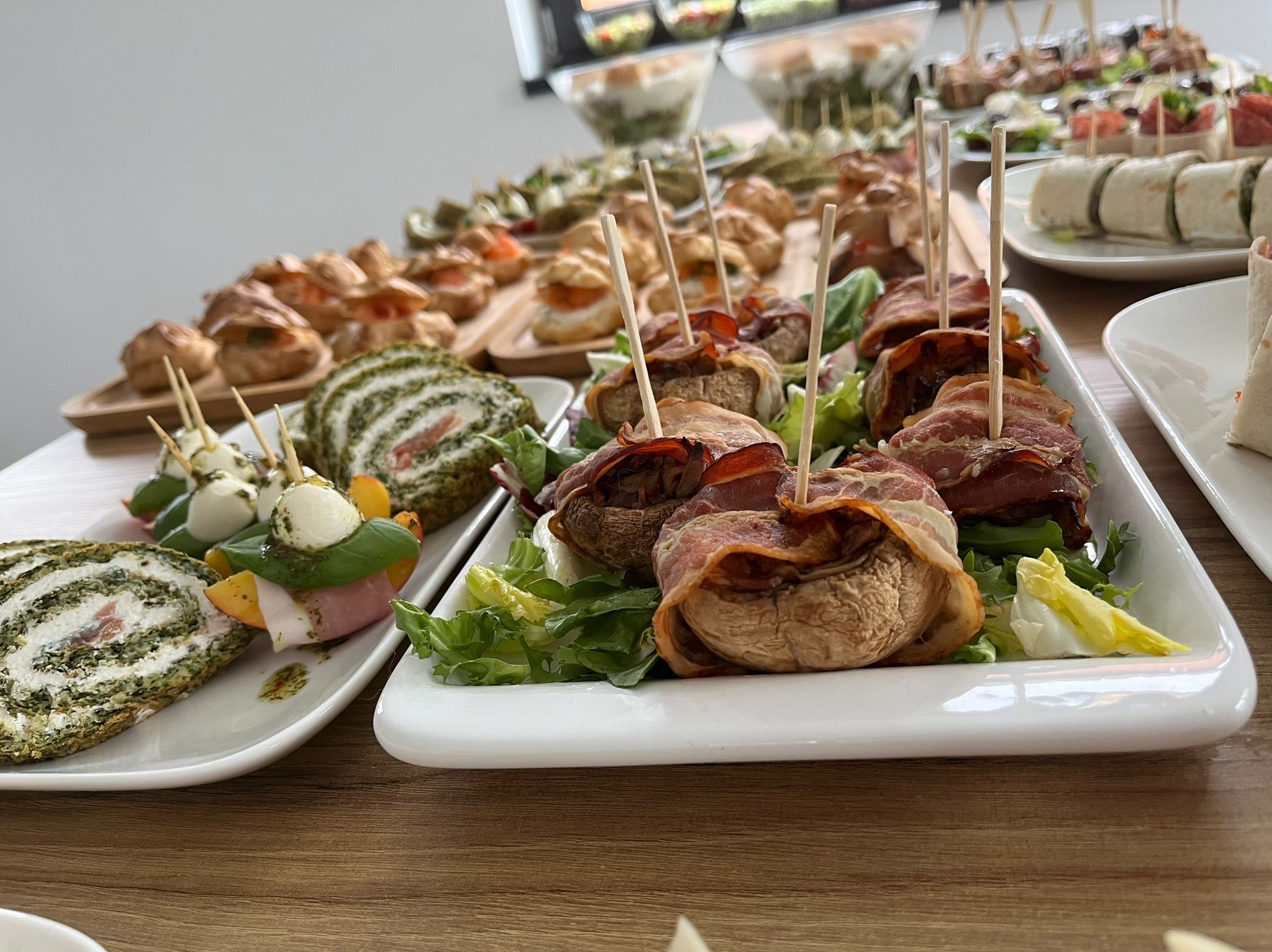 Dekoracyjnie ułożone przekąski na drewnianym stole: roladki szpinakowe, koreczki z mozzarelli i papryki, pieczarki owinięte boczkiem na sałacie, tortille z farszem.