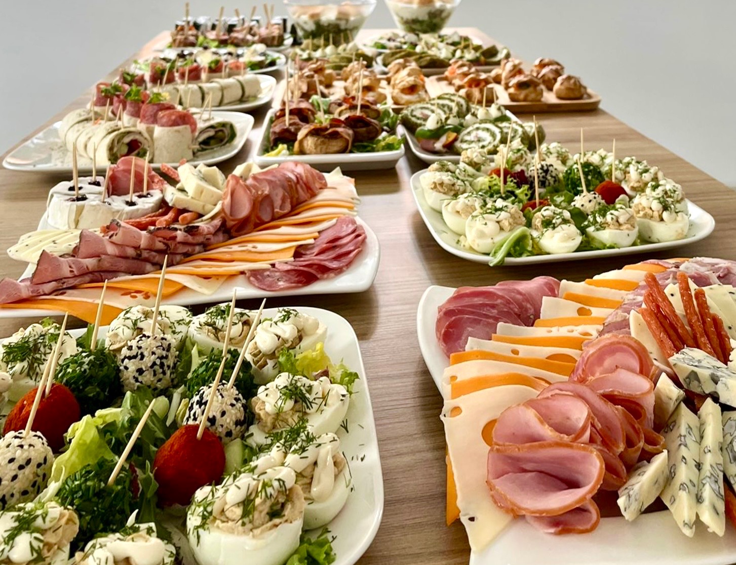 Stół z różnorodnymi przekąskami: roladki z tortilli, koreczki z grzybami, jajka faszerowane, deski wędlin i serów, wszystko na białych talerzach.