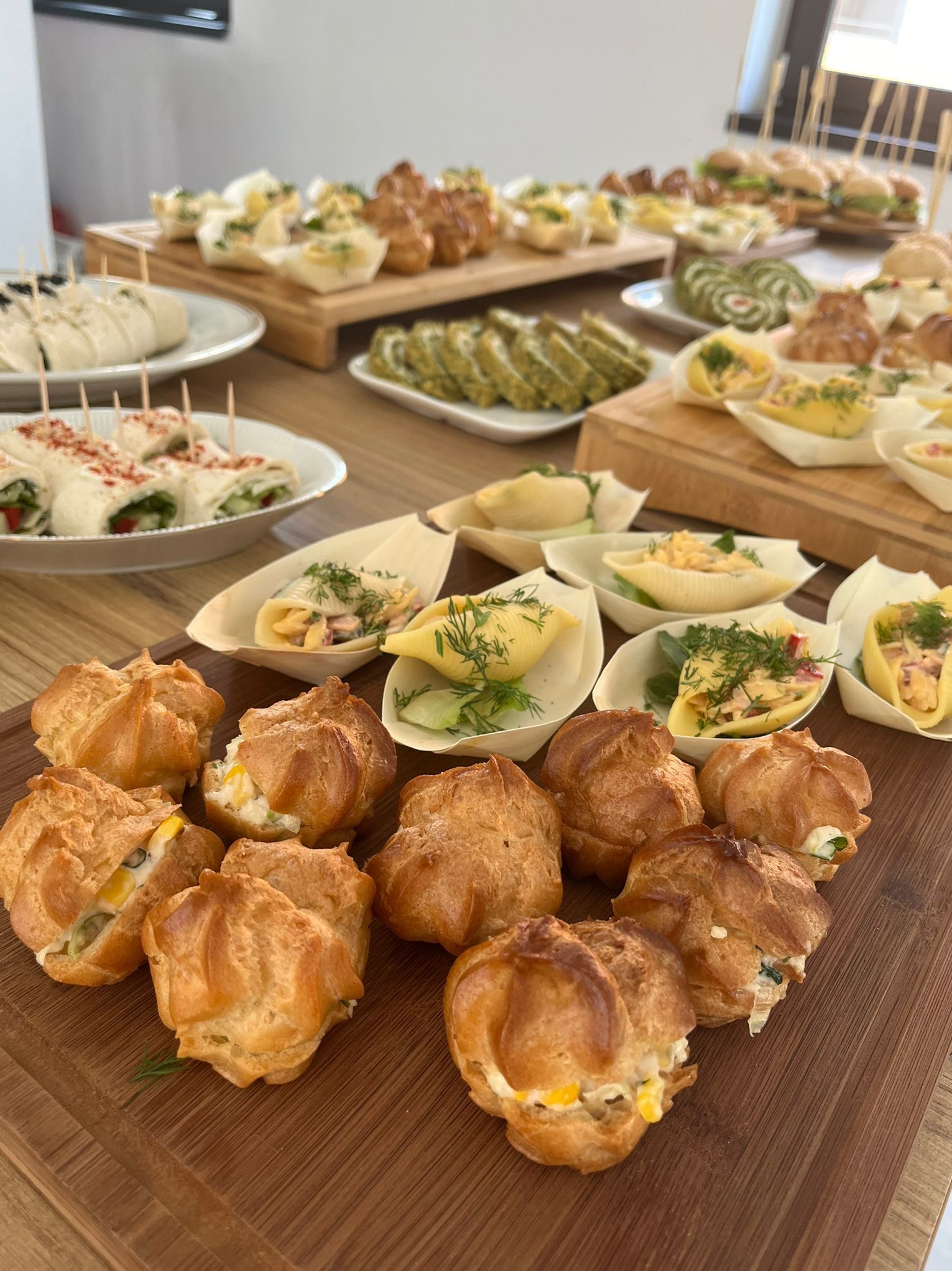 Różnorodne przekąski na drewnianych deskach i talerzach: mini-kanapeczki, roladki z warzywami i kremem, ptysie z nadzieniem, makaronowe muszle z sałatką, wszystko ułożone na stole.
