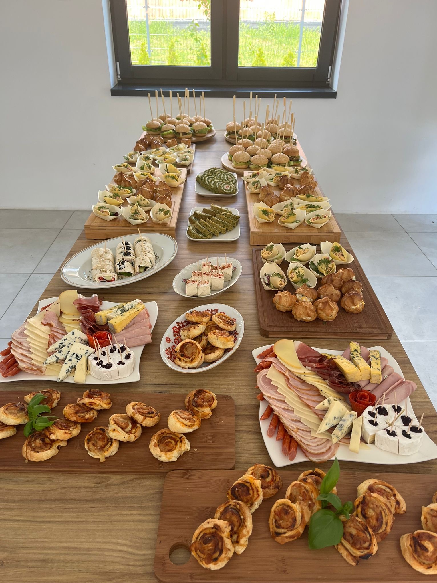 Stół z różnorodnymi przekąskami: mini burgery na wykałaczkach, roladki z szynką, tartaletki z farszem, paszteciki, koreczki serowo-wędliniarskie na deskach i talerzach, ustawione pod oknem.