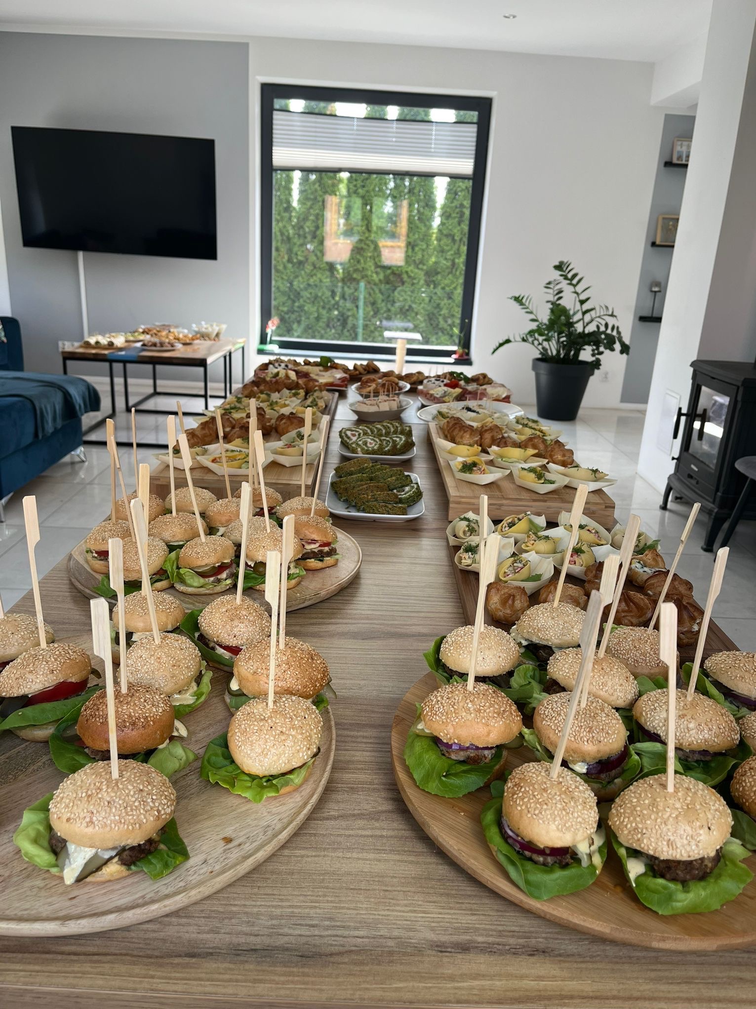 Stół z różnorodnymi przekąskami: mini burgery z wykałaczkami, croissanty, sałatki w małych naczyniach i zielone roladki, przygotowane w jasnym wnętrzu z dużym oknem.