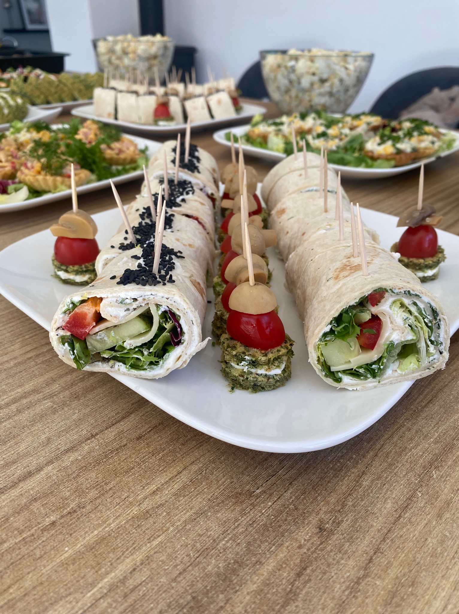 Tortilla wrap przekrojona na kawałki, spięta wykałaczkami, obok mini-kanapeczki z pomidorkiem i pieczarką na drewnianym stole, w tle inne przekąski.
