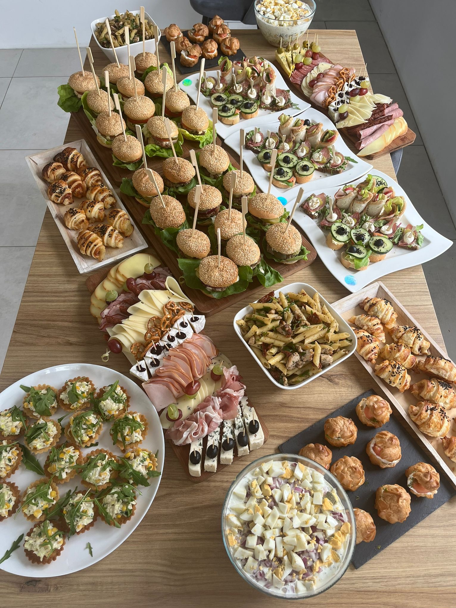 Stół z różnorodnym cateringiem: mini burgery z sezamem na wykałaczkach, tartaletki z sałatką, kanapeczki, croissanty z czarnuszką, sałatka jarzynowa i deska serów z wędlinami, całość ułożona...