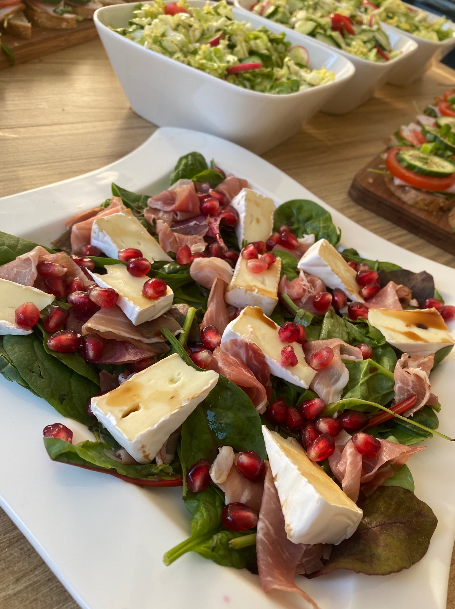 Sałatka ze szpinaku, szynki parmeńskiej, sera brie i granatu na białym talerzu, z sałatkami w tle.