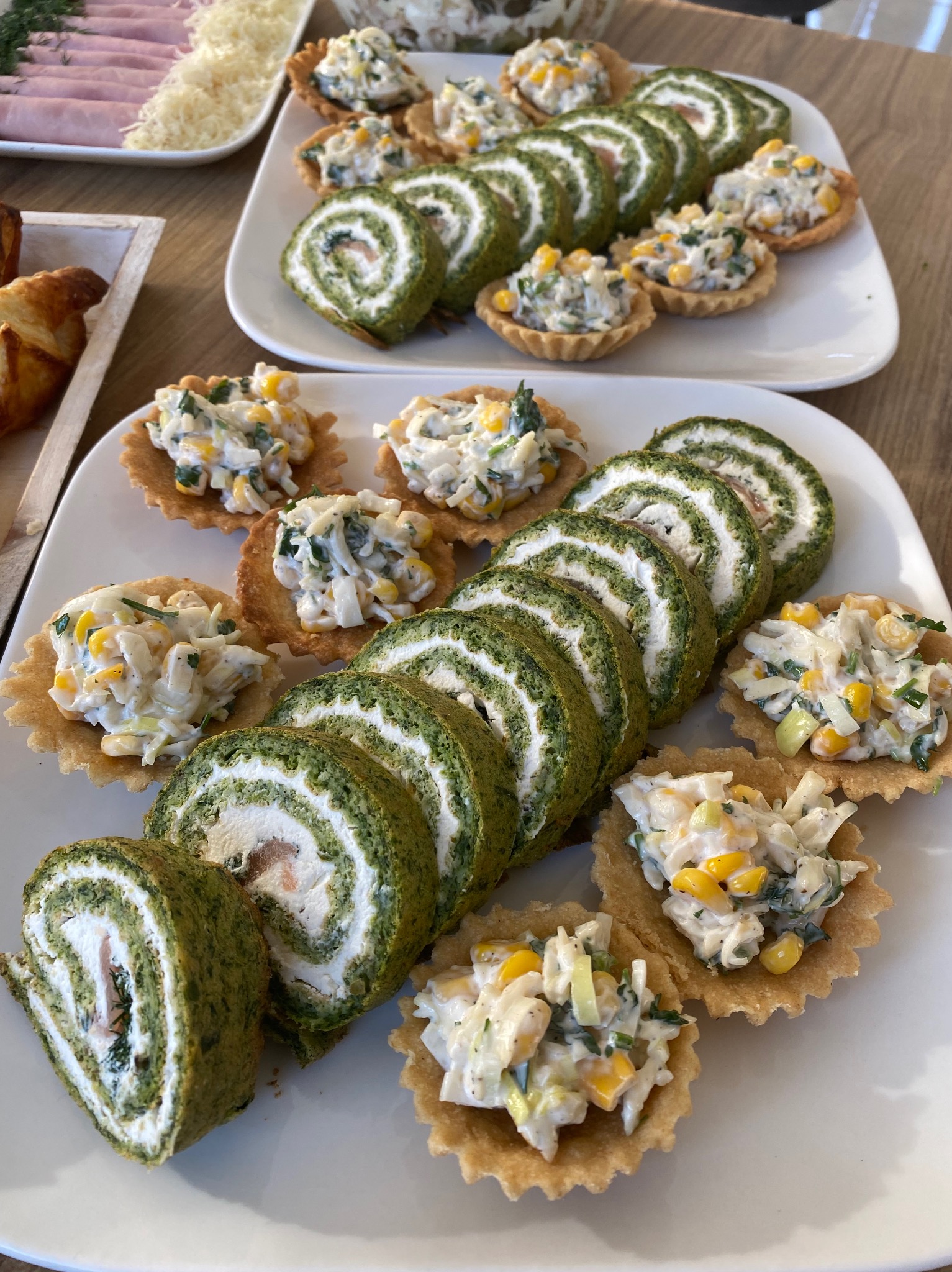 Dwa białe talerze z roladkami szpinakowymi z łososiem i tartaletkami z sałatką warzywną, ułożone na drewnianym stole, widoczne również wędliny i ciastka.