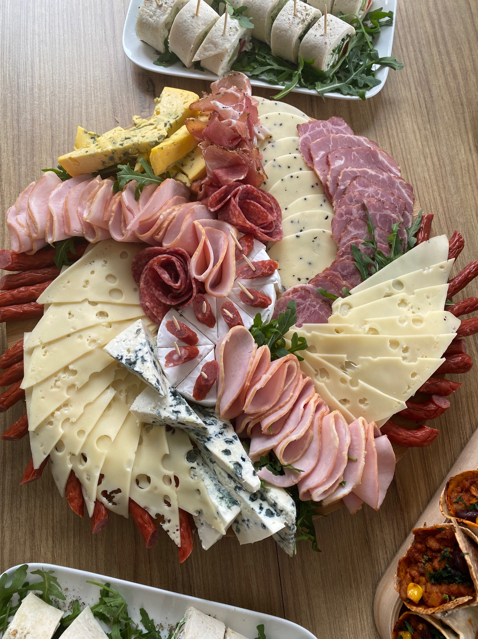 Dekoracyjna deska z różnorodnymi serami, wędlinami i roladkami na jasnym drewnianym stole, uzupełniona świeżą rukolą i przekąskami na talerzach.