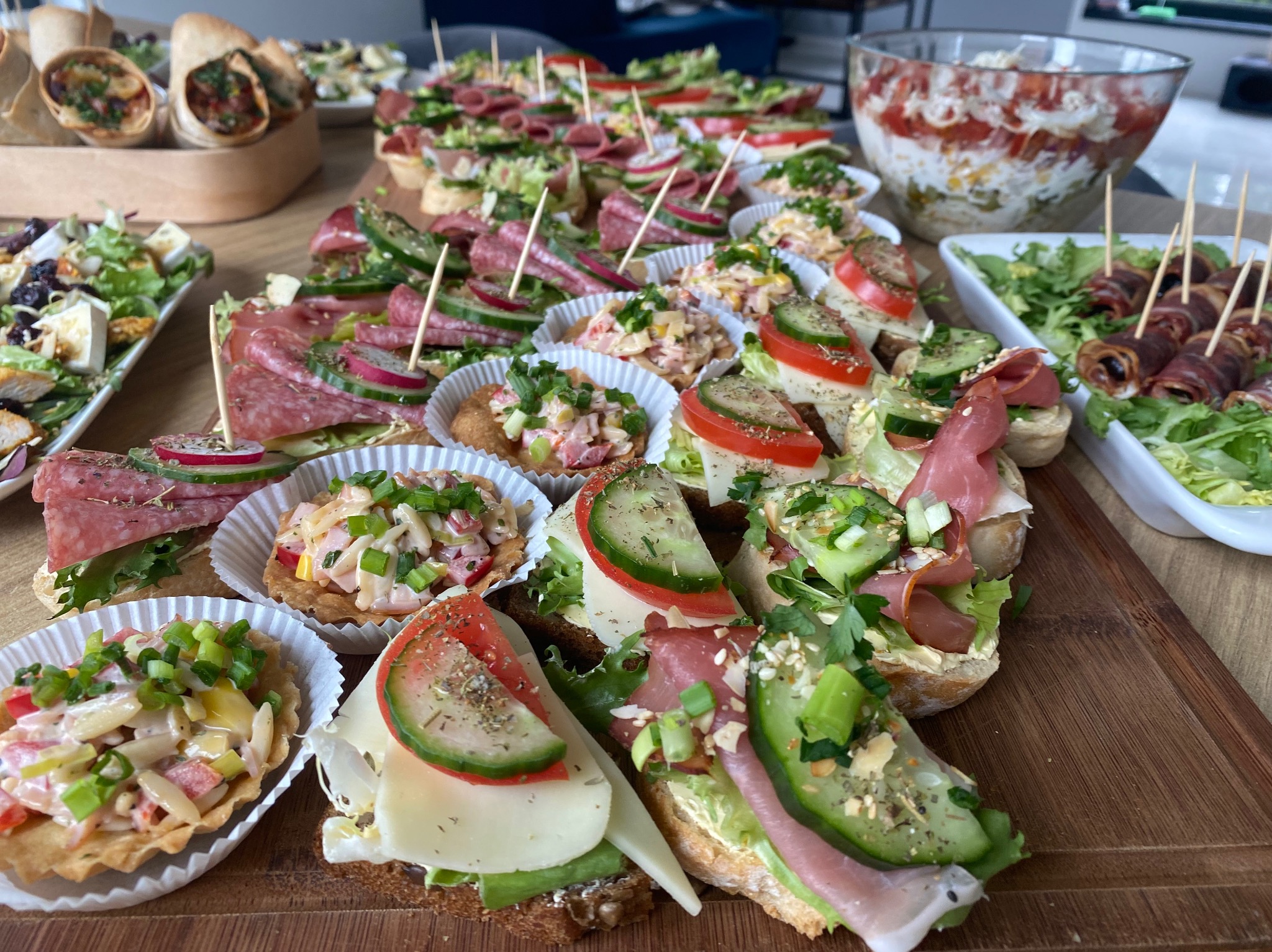 Różnorodny wybór kanapek i przekąsek na drewnianej desce, w tym zawijane tortille, sałatki, koreczki z salami i ogórkiem, oraz tartaletki z sałatką, wszystko przygotowane na przyjęcie.