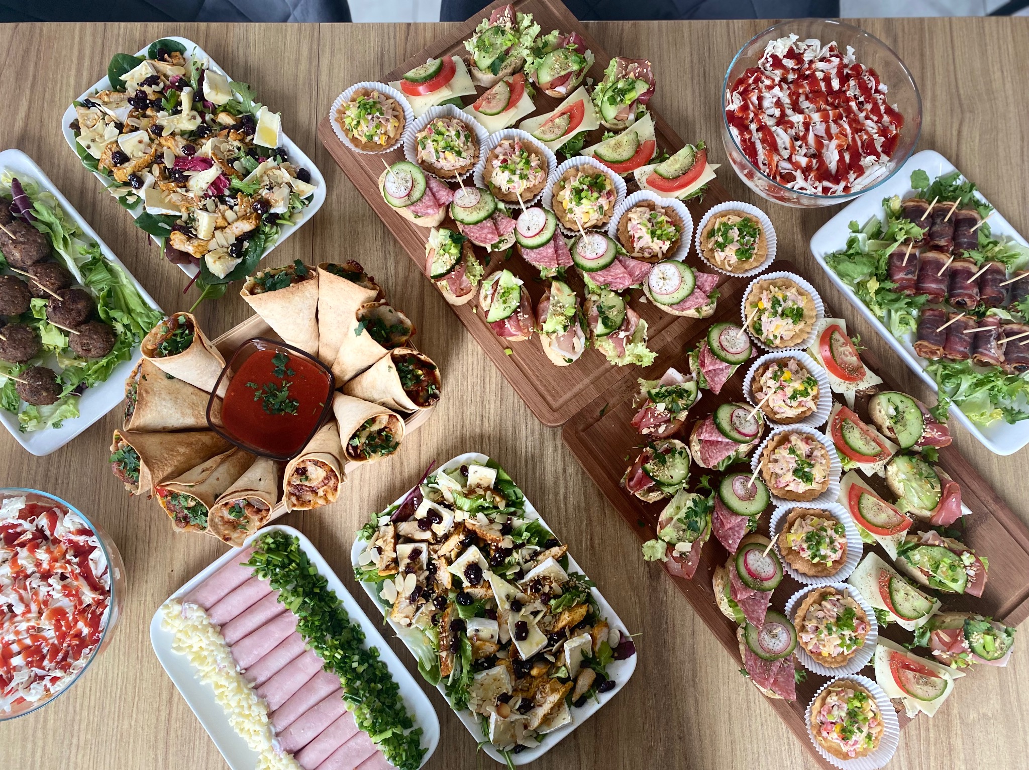 Stół zastawiony różnorodnymi przekąskami: tortille z sosem, koreczki z szynki parmeńskiej, sałatki z serem camembert i kurczakiem, mini-kanapeczki z salami i ogórkiem, miska sałatki z sosem...