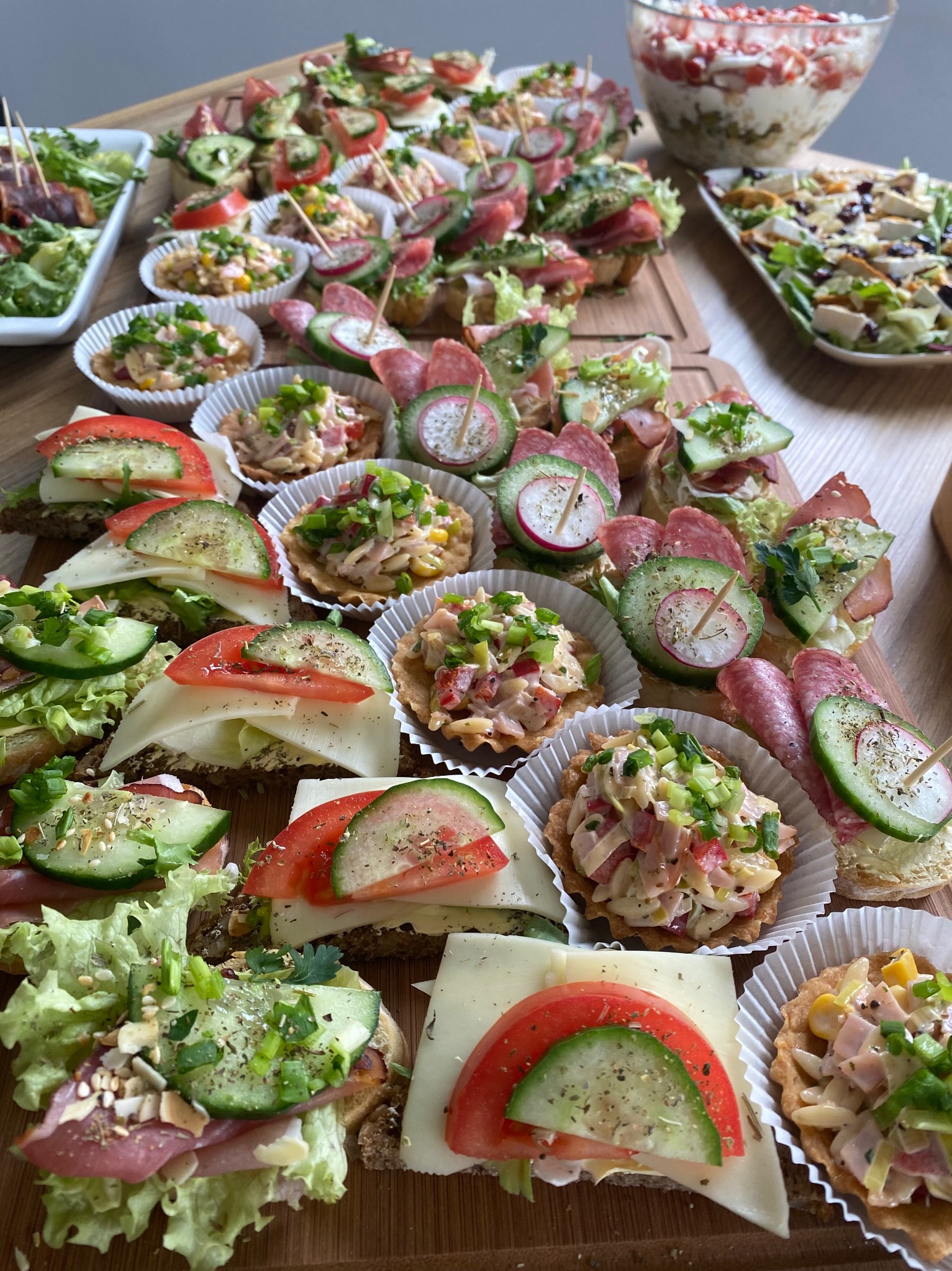Różnorodne kanapki i tartaletki na drewnianej desce, udekorowane świeżymi warzywami, wędlinami i ziołami, tworzące apetyczną kompozycję przekąsek.