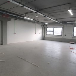 PC INSTAL - Wnętrze hali produkcyjnej w trakcie prac instalacyjnych: widoczne koryta kablowe pod sufitem, punkty wyprowadzenia przewodów na ścianie, surowa posadzka.