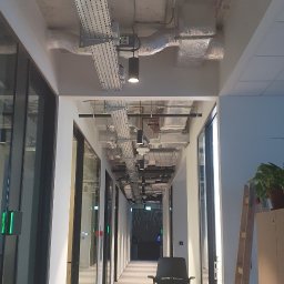 PC INSTAL - Długi korytarz biurowy z widocznymi instalacjami pod sufitem, w tym korytami kablowymi i systemem wentylacji, z krzesłem biurowym i odkurzaczem na podłodze.