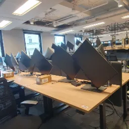 Rząd biurek z monitorami w nowym biurze, widoczne kable i niezapakowane akcesoria, oświetlenie sufitowe włączone.