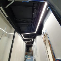 PC INSTAL - Długi korytarz biurowy w trakcie wykańczania, z zamontowanym nowoczesnym oświetleniem liniowym LED w kształcie kwadratów pod sufitem i widocznymi elementami instalacji pod sufitem. Ściany w trakcie...