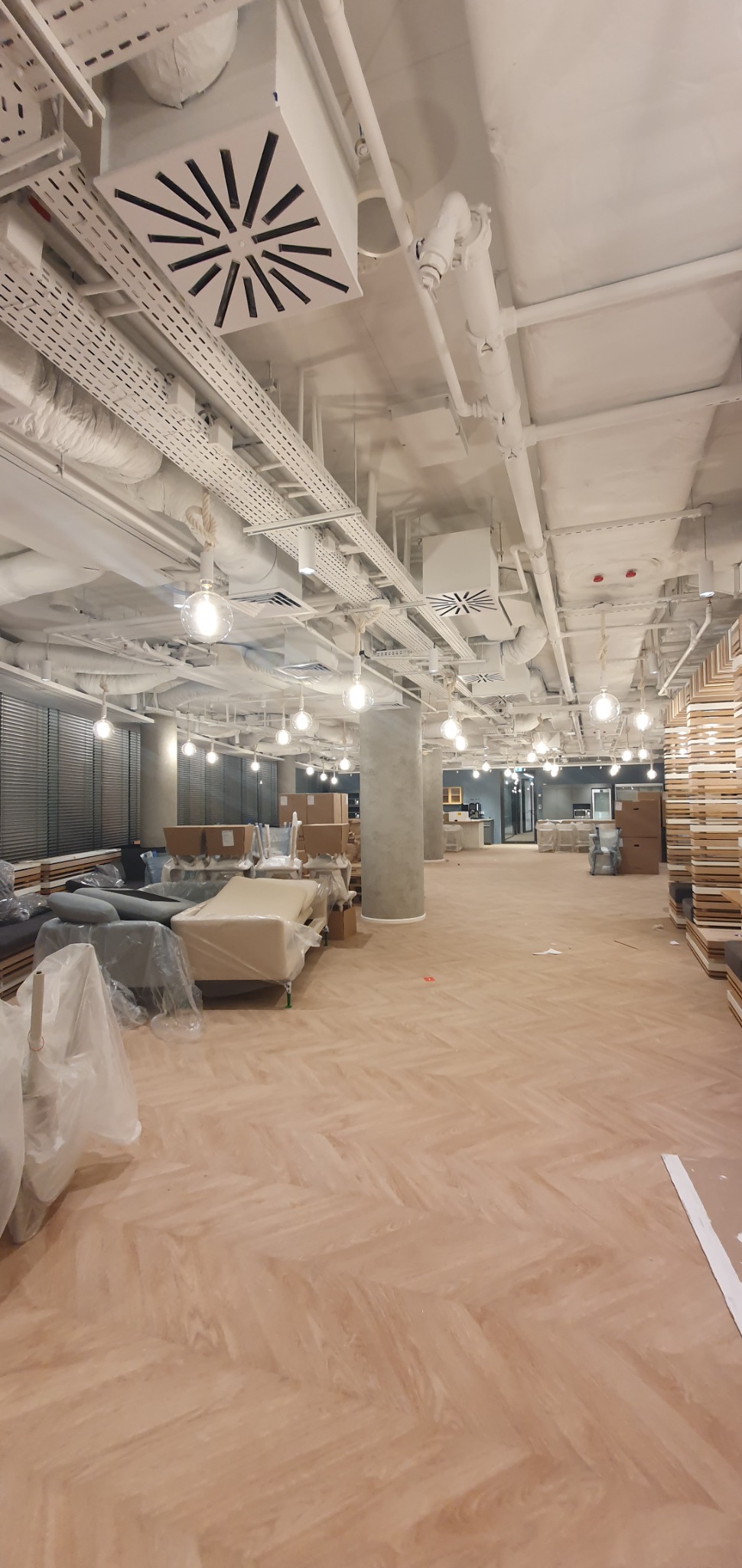 Wnętrze biurowe w trakcie wykańczania, widoczne elementy wentylacji pod sufitem, oświetlenie w postaci wiszących kul oraz meble zabezpieczone folią