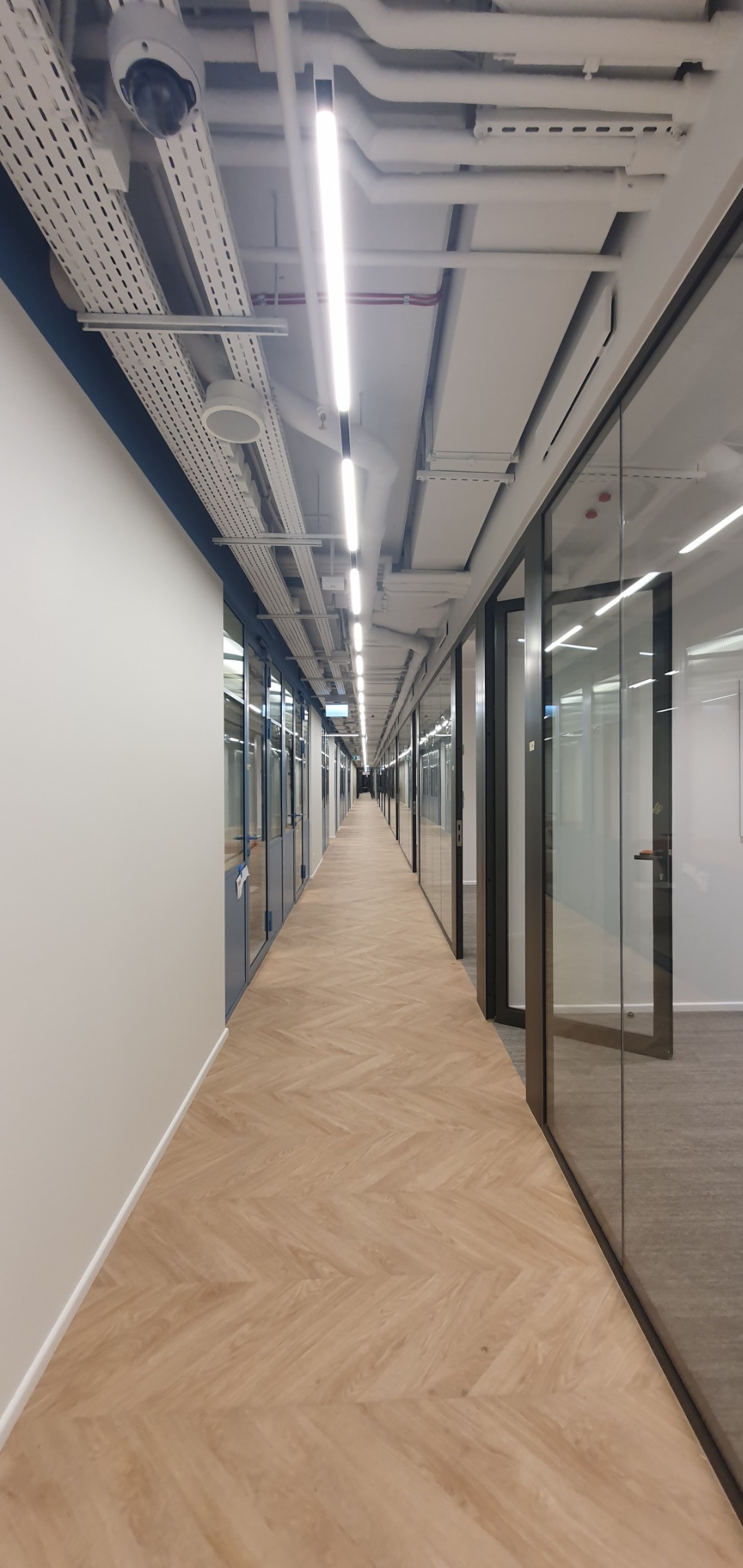 Długi korytarz biurowy z parkietem w jodełkę, oświetlony liniowymi lampami LED, z widocznymi instalacjami pod sufitem i kamerą monitoringu.
