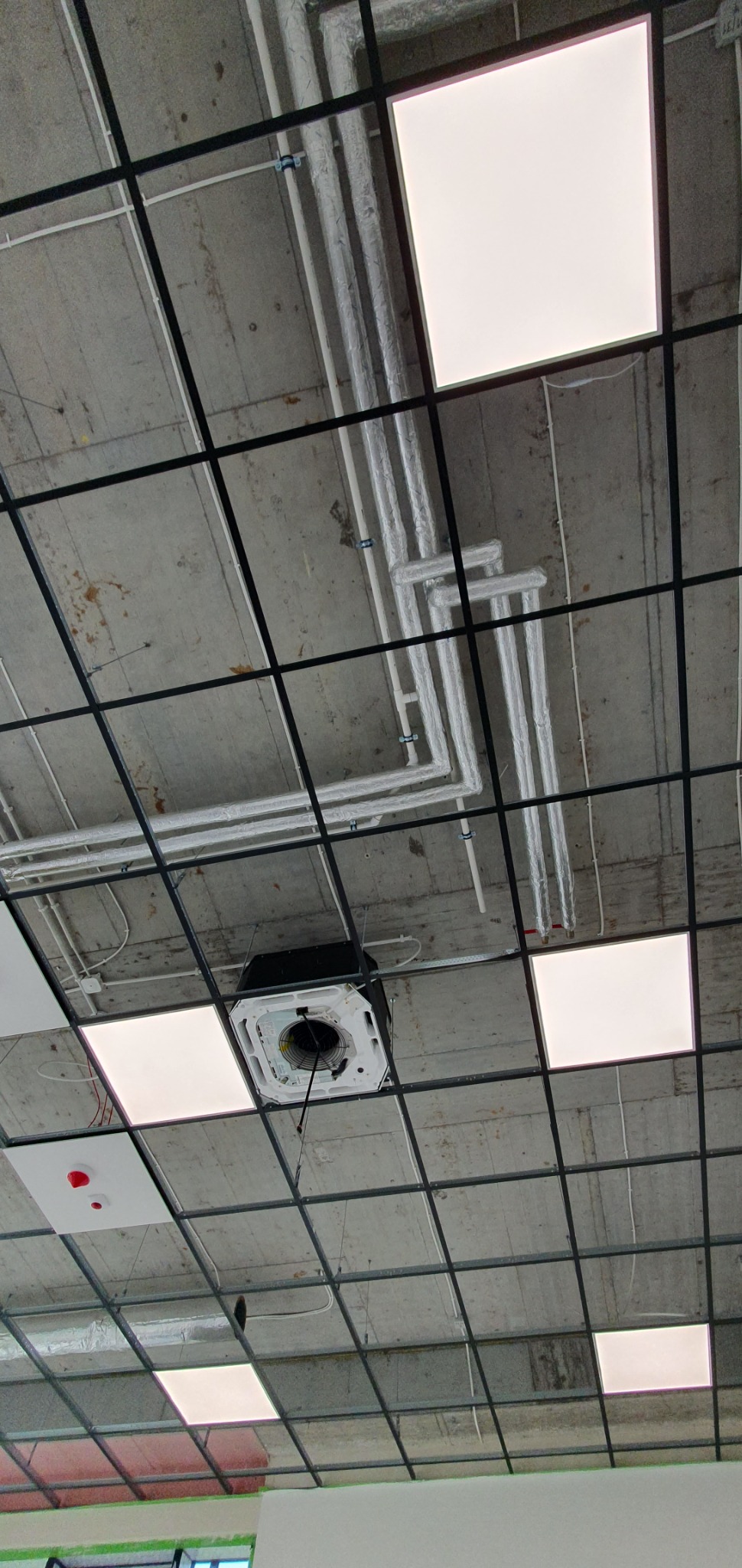 Widok sufitu podwieszanego z zainstalowanymi panelami LED, systemem rur izolowanych termicznie i klimatyzatorem kasetonowym z widocznymi przewodami.