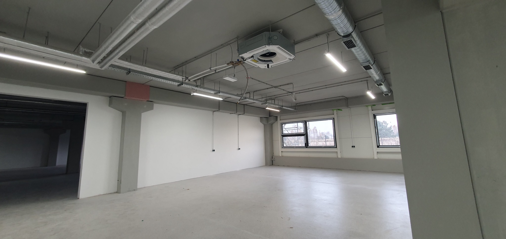 Wnętrze hali przemysłowej z widocznym systemem wentylacji, klimatyzacji i oświetlenia liniowego pod sufitem. Dwa okna z czarnymi ramami oświetlają przestrzeń.