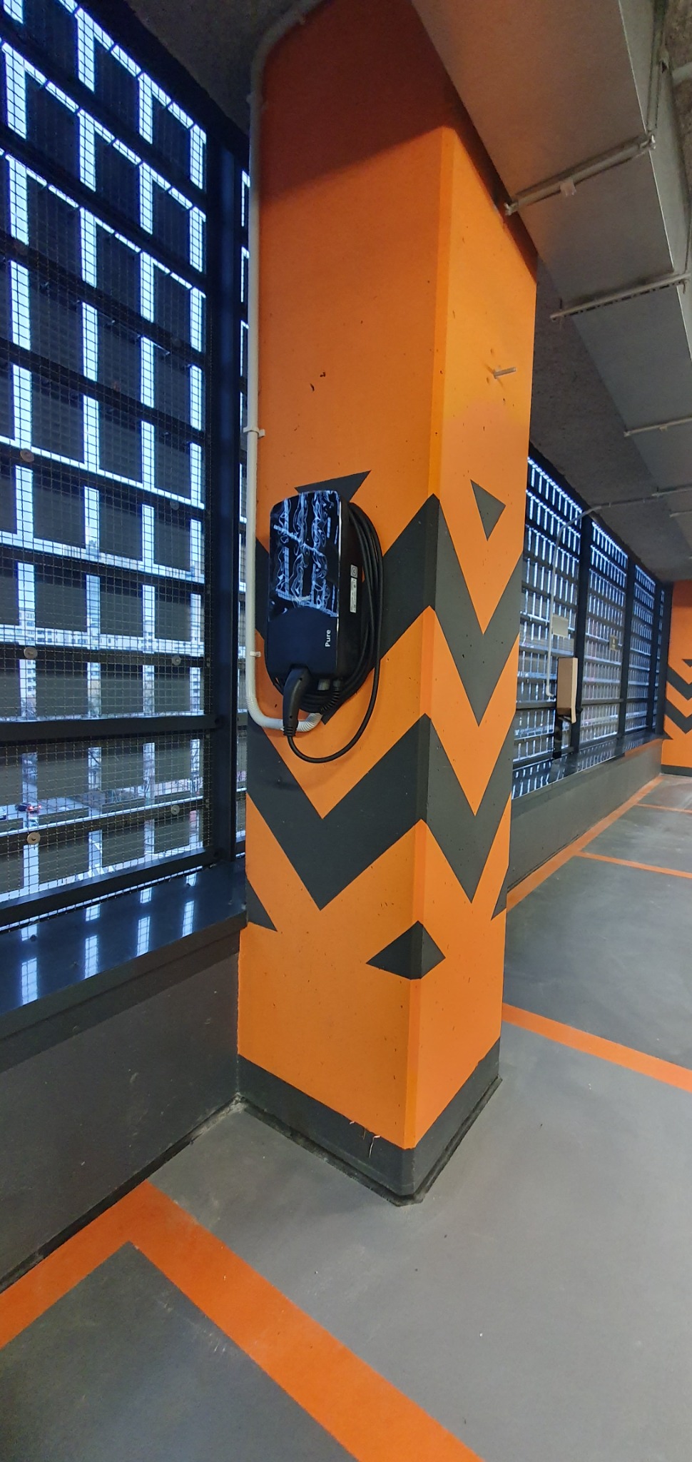 Stacja ładowania pojazdów elektrycznych marki Pure zamontowana na pomarańczowym filarze z szarymi chevronami w garażu podziemnym.
