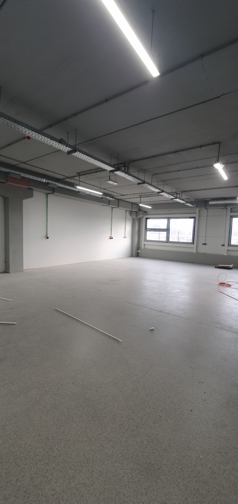 Wnętrze hali produkcyjnej w trakcie prac instalacyjnych: widoczne koryta kablowe pod sufitem, punkty wyprowadzenia przewodów na ścianie, surowa posadzka.