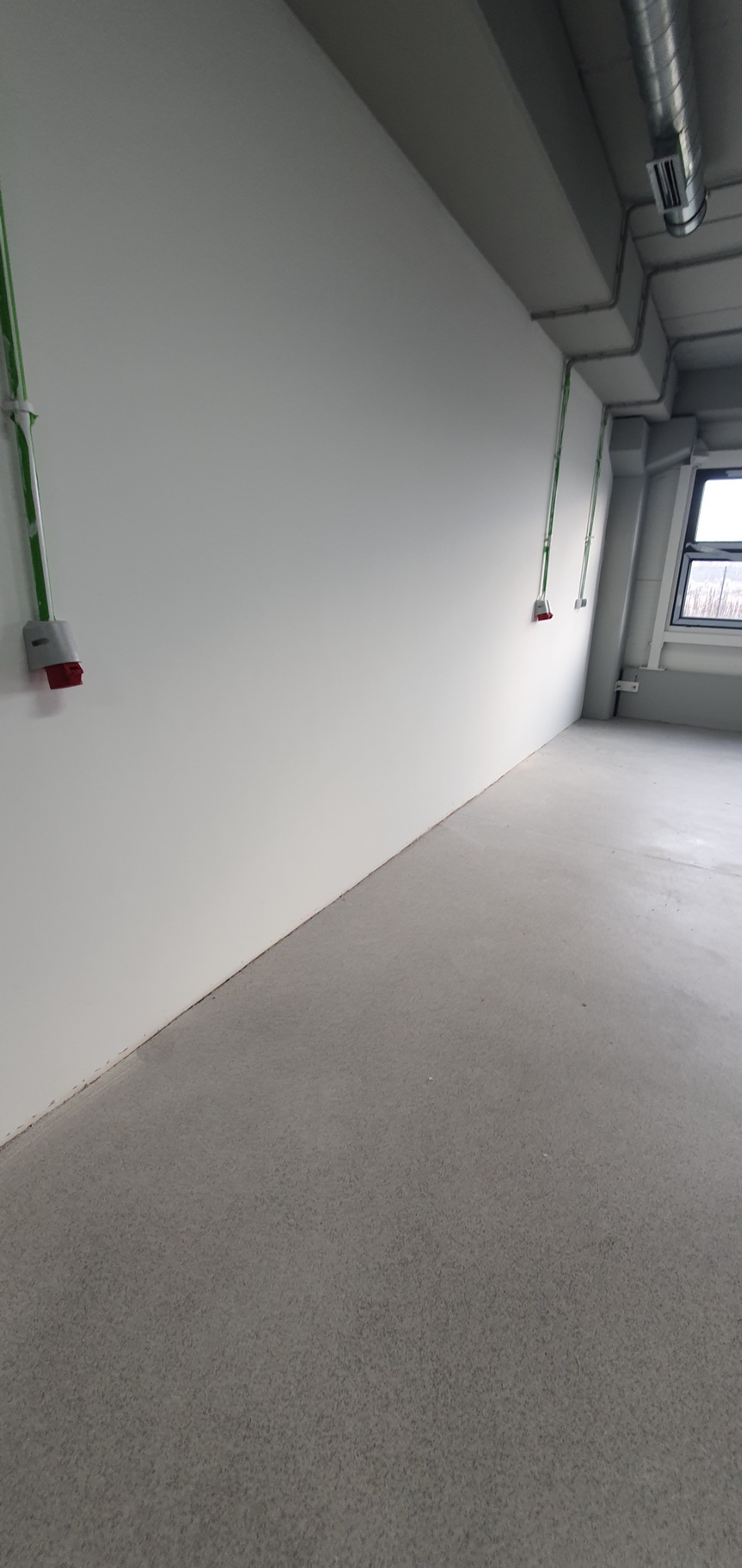 Puste pomieszczenie z szarą podłogą i białymi ścianami, widoczne gniazda elektryczne w szarych obudowach z czerwonymi wtykami, poprowadzone po ścianie w zielonych rurkach, oraz rury wentylacyjne...