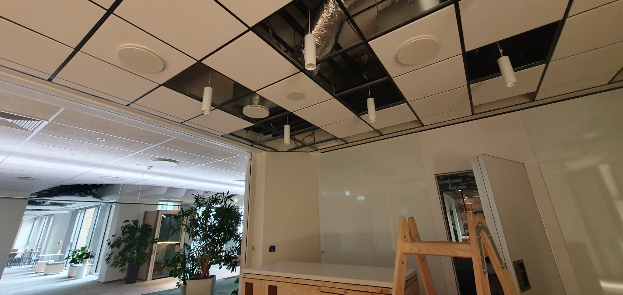 Widok na sufit podwieszany z zainstalowanymi okrągłymi lampami i cylindrycznymi oprawami oświetleniowymi, część paneli zdjęta, widoczne elementy konstrukcyjne, w tle widok na nowoczesne biuro...