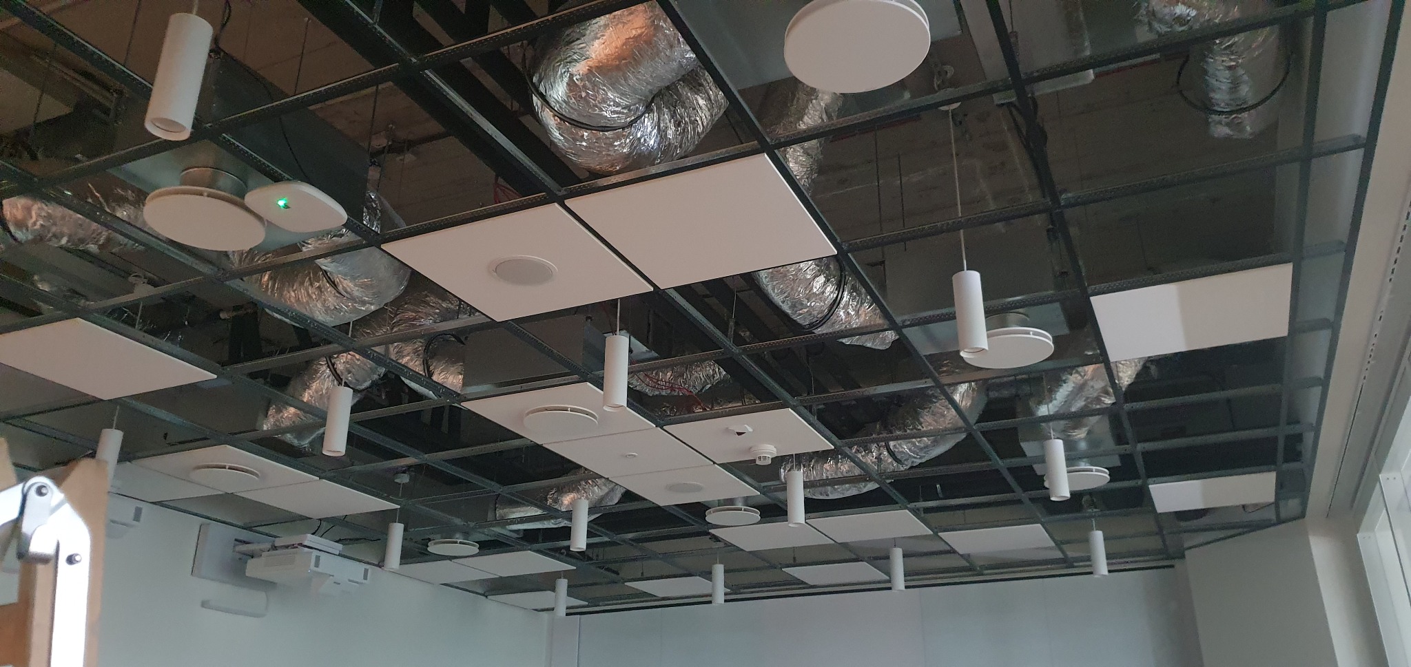 Sufit podwieszany rastrowy z widocznymi instalacjami wentylacyjnymi, oświetleniem cylindrycznym i okrągłymi głośnikami, oraz detektorami dymu.