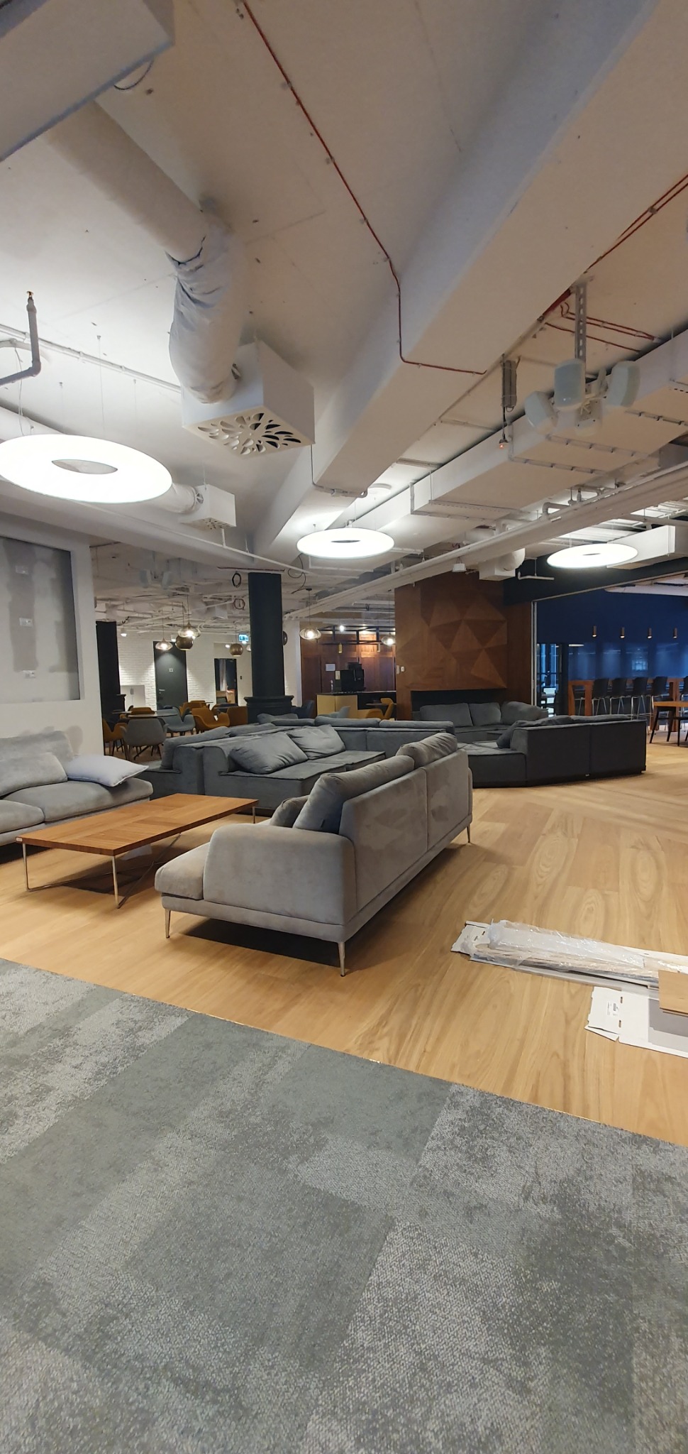 Przestronne biuro z szarymi sofami modułowymi, drewnianym parkietem i okrągłymi lampami sufitowymi, widoczne elementy instalacji elektrycznej pod sufitem.