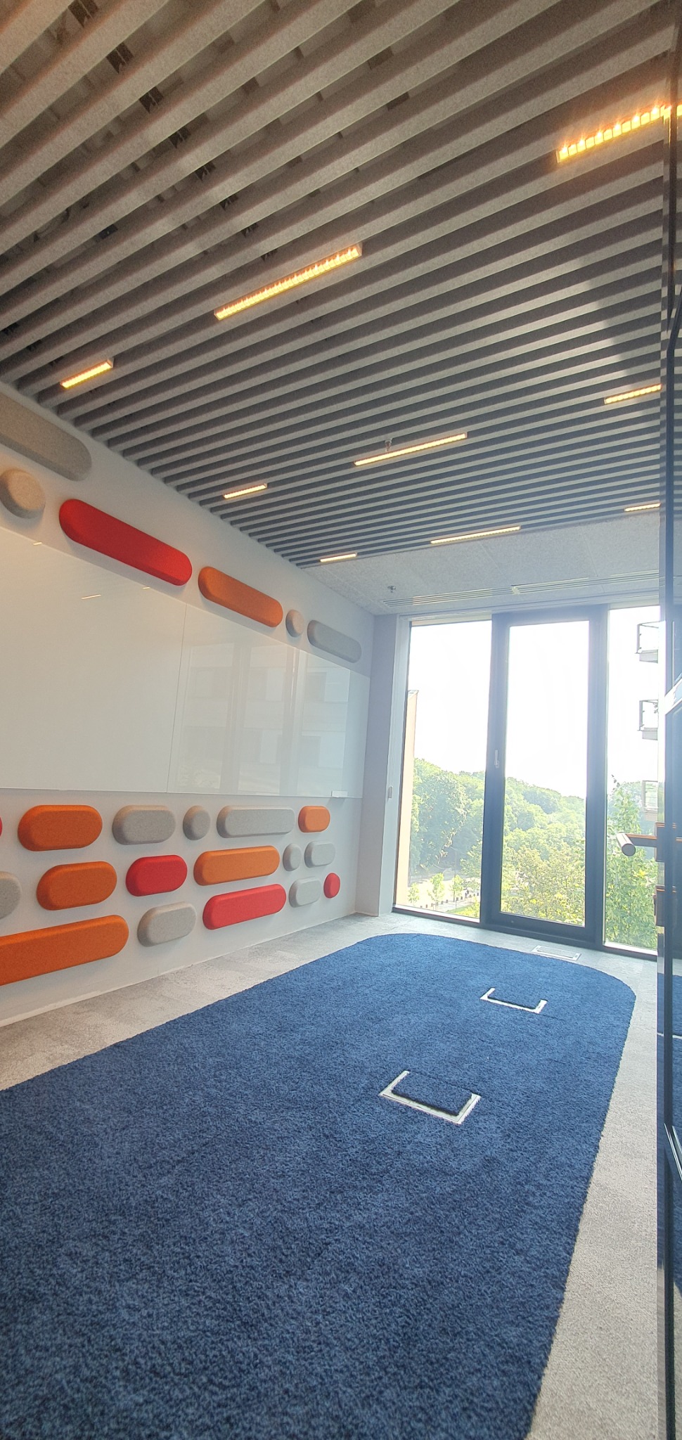 Nowoczesne biuro z akustycznym sufitem lamelowym, liniowym oświetleniem LED, białą tablicą z kolorowymi panelami akustycznymi i niebieską wykładziną dywanową z ukrytymi gniazdami podłogowymi.