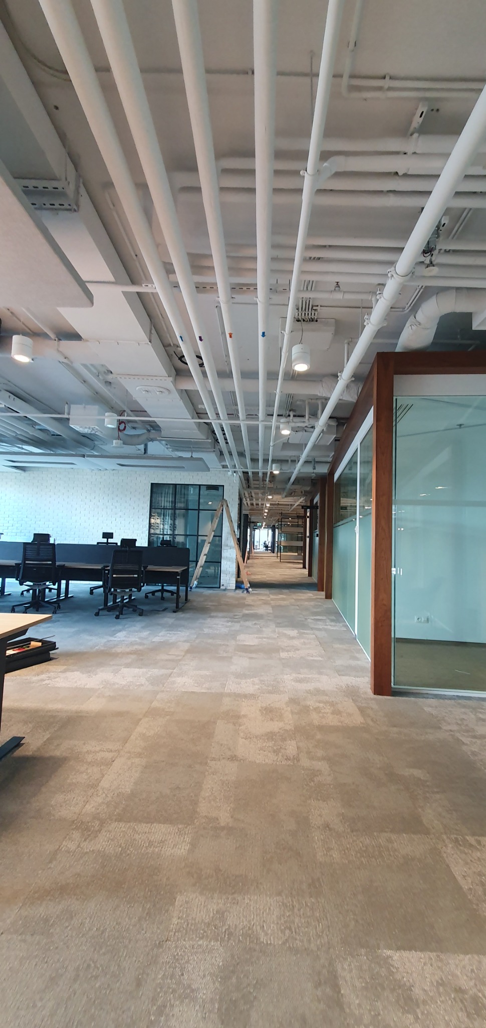 Wnętrze biurowe z widocznymi instalacjami pod sufitem, drabina oparta o ścianę, oświetlenie sufitowe, wykładzina dywanowa w odcieniach szarości.