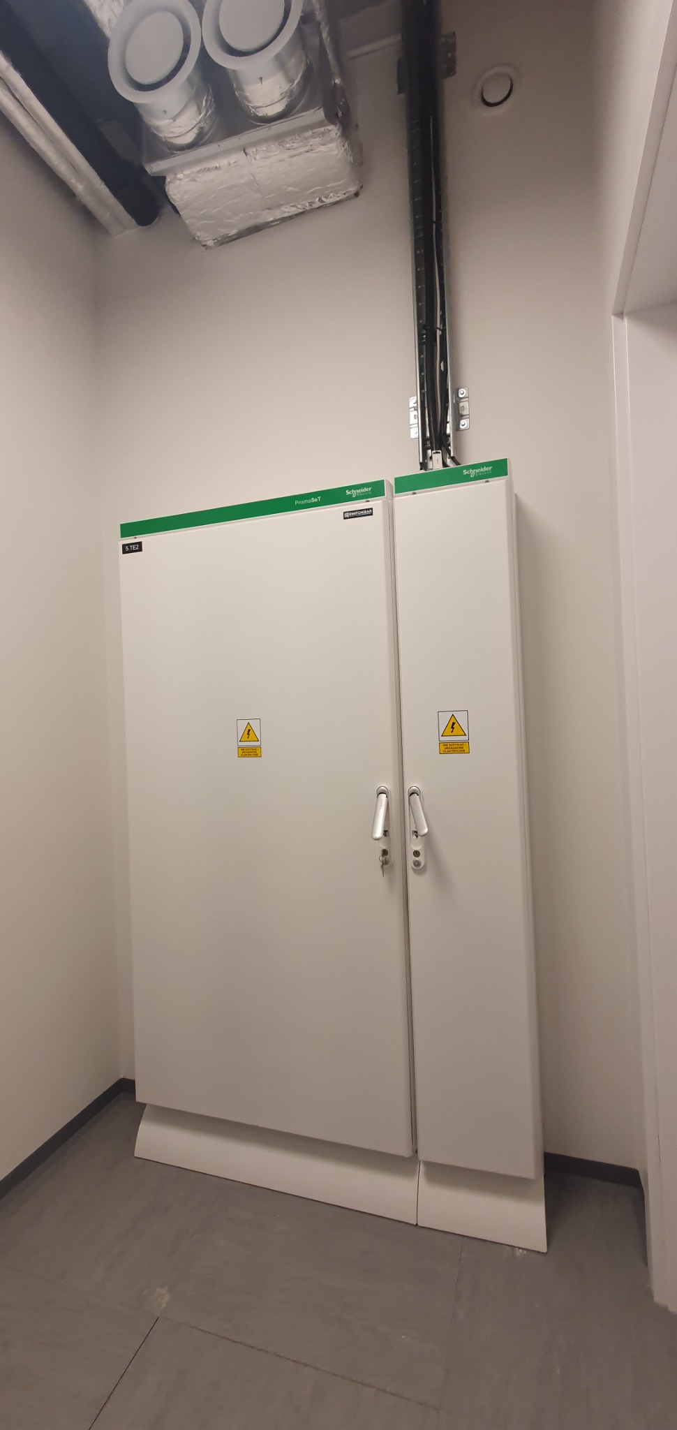 Biała szafa elektryczna z dwoma drzwiami, ostrzeżeniami o napięciu i logo Schneider Electric, zamontowana w pomieszczeniu z szarą podłogą i widocznymi przewodami w górnej części ściany.