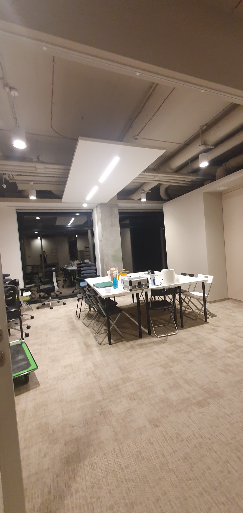 Przestronne biuro z białymi stołami, krzesłami i widocznymi przewodami elektrycznymi pod sufitem, oświetlone lampami LED.