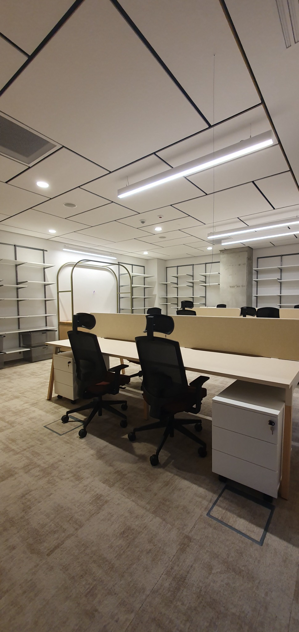 Wnętrze biura z rzędami biurek i krzeseł, oświetlone liniowymi lampami sufitowymi. Widoczne puste regały w tle.