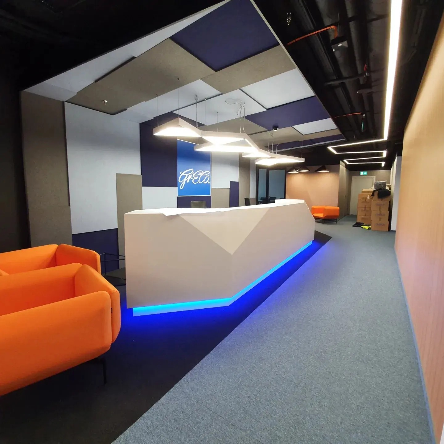 Nowoczesne biuro z recepcją podświetloną na niebiesko, pomarańczowe sofy, designerskie oświetlenie sufitowe i panele akustyczne na ścianach.