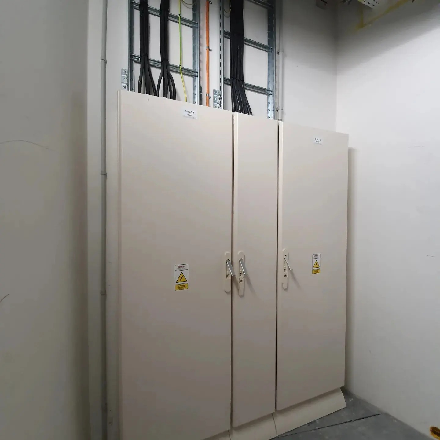 Trzy białe szafy elektryczne z uchwytami i naklejkami ostrzegawczymi, zamontowane na ścianie w pomieszczeniu z widocznymi kablami w korytkach nad nimi.