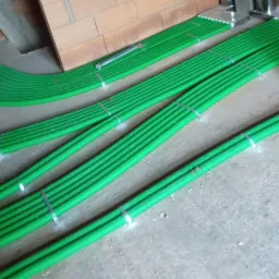 Zielone, karbowane rury wentylacyjne ułożone równolegle na podłodze podczas instalacji, zabezpieczone metalowymi obejmami, widoczne fragmenty ceglanej ściany i surowego betonu.