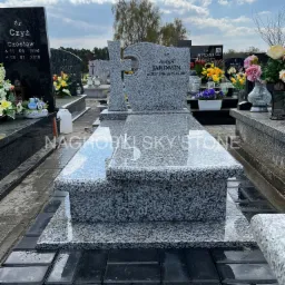 Granitowy pomnik z krzyżem i wygrawerowanymi danymi zmarłej osoby, umieszczony na cmentarzu, z nagrobkami w tle i refleksami światła na polerowanej powierzchni.