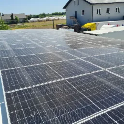 Rozległa instalacja fotowoltaiczna na dachu budynku przemysłowego, z widocznymi modułami i konstrukcją mocującą. W tle budynek biurowy z żółtą ciężarówką dostawczą i zielone pola uprawne...