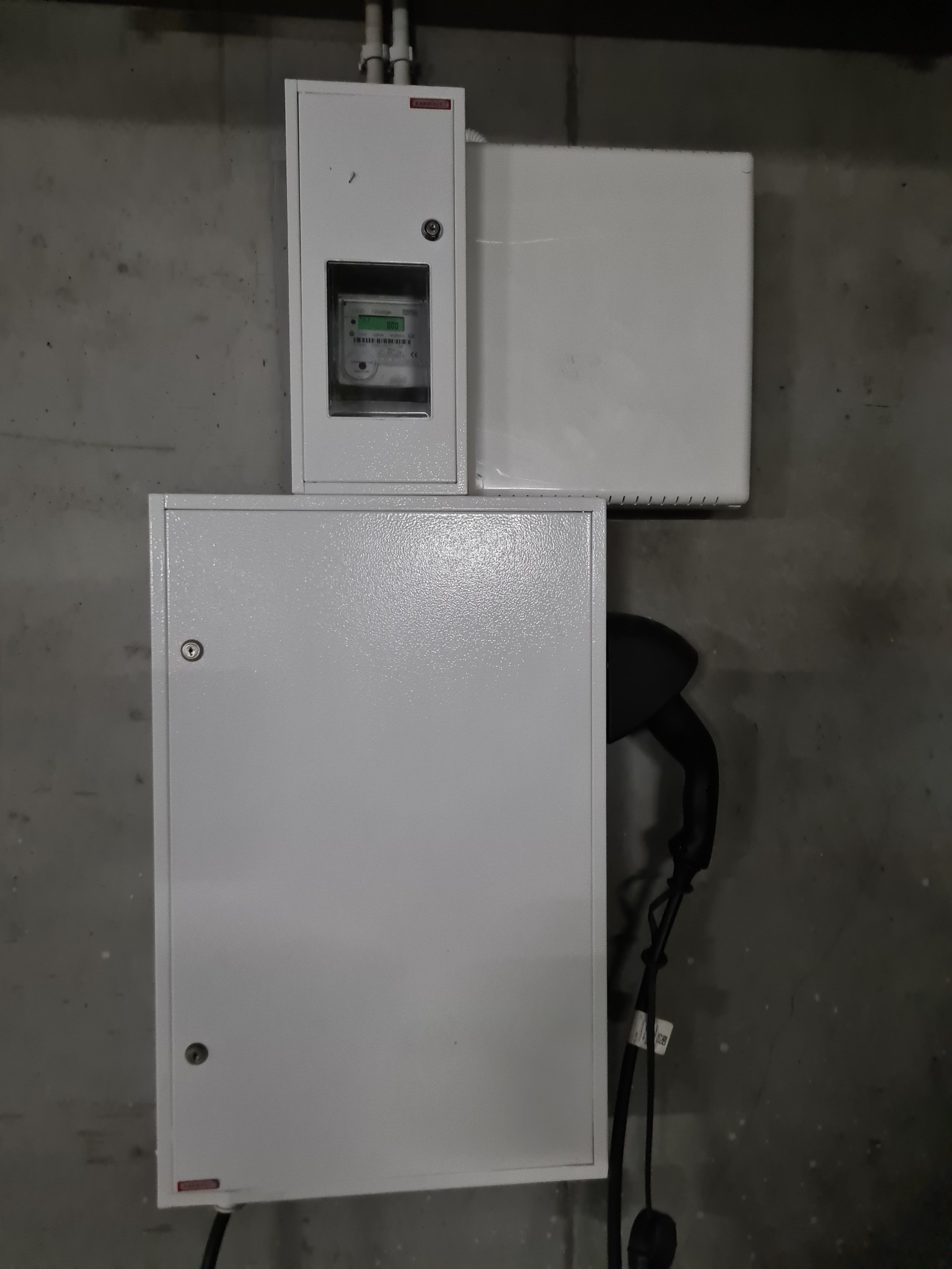 Biała ładowarka 11 kW do samochodu elektrycznego zamontowana na szarej, betonowej ścianie w garażu wielostanowiskowym, obok znajduje się licznik energii w białej obudowie i biała skrzynka elektryczna.