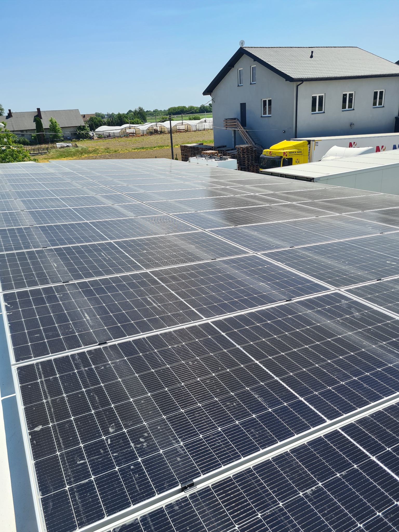 Rozległa instalacja fotowoltaiczna na dachu budynku przemysłowego, z widocznymi modułami i konstrukcją mocującą. W tle budynek biurowy z żółtą ciężarówką dostawczą i zielone pola uprawne...