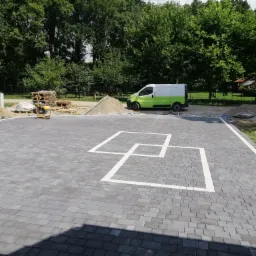 Ułożona szara kostka brukowa z białymi kwadratami dekoracyjnymi, w tle zielony bus, palety z kostką i ubijarka wibracyjna.