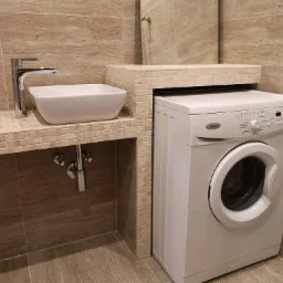 Biała, prostokątna umywalka nablatowa z chromowaną baterią, obok pralka w łazience wykończonej beżowymi płytkami imitującymi trawertyn, z mozaikowymi akcentami.