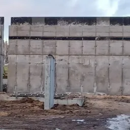 Wznoszenie żelbetowej konstrukcji ścian budynku z widocznymi elementami szalunkowymi i zbrojeniowymi, w tle fragment domu i drzewa.