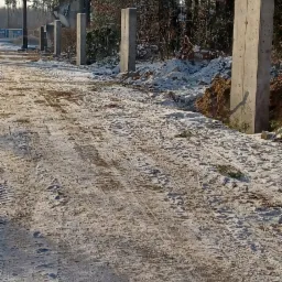 Betonowe słupki ustawione wzdłuż drogi gruntowej pokrytej śniegiem, w tle las i pomnik.