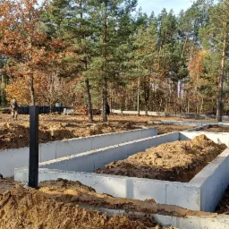 Wylane betonowe fundamenty pod budowę domu jednorodzinnego na zalesionej działce, widoczne czarne słupki i częściowo zasypane ziemią zagłębienia fundamentowe.