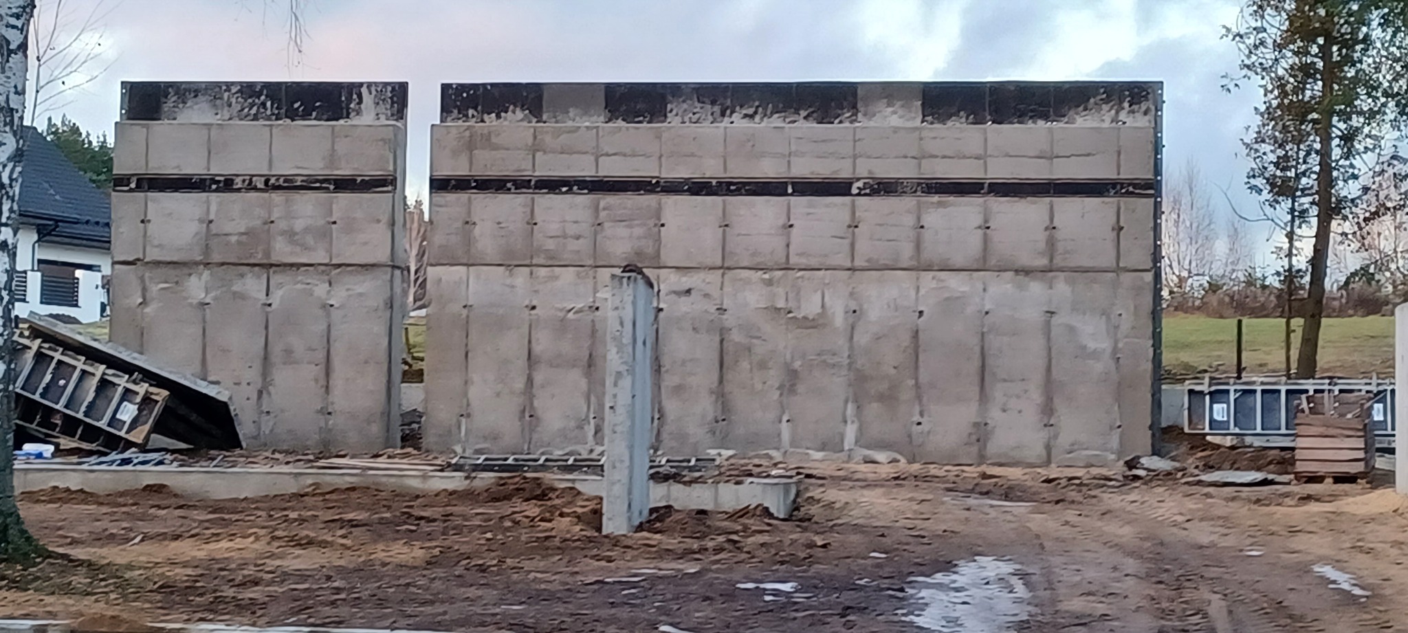 Wznoszenie żelbetowej konstrukcji ścian budynku z widocznymi elementami szalunkowymi i zbrojeniowymi, w tle fragment domu i drzewa.