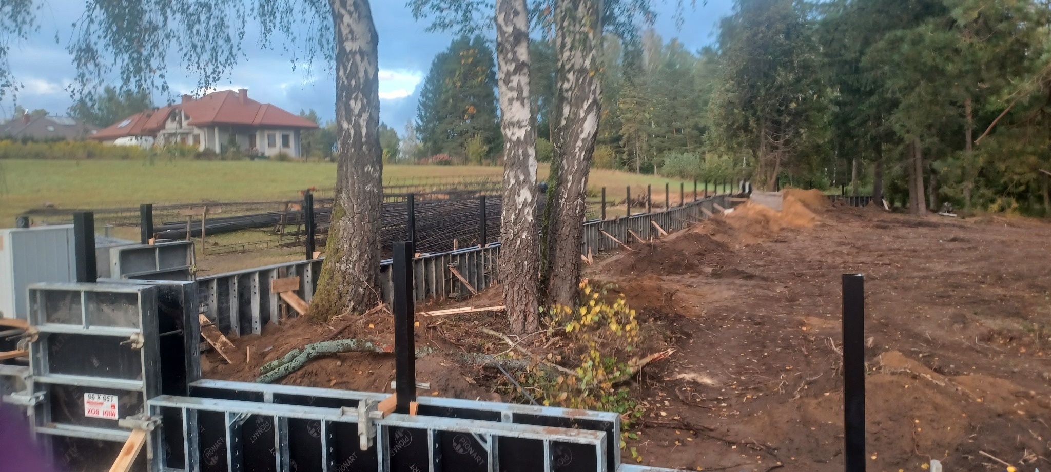 Budowa fundamentów: Szalunki zbrojeniowe w wykopie, na tle drzew i domu z czerwoną dachówką.