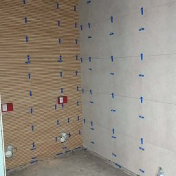 Marcin Domagalski. Usługi budowlane DOM-BUD - Ściana z ułożonymi płytkami imitującymi drewno i jasnymi płytkami ceramicznymi, zabezpieczonymi niebieską taśmą. Widoczne instalacje hydrauliczne i poziomowanie laserowe.