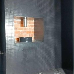 Marcin Domagalski. Usługi budowlane DOM-BUD - Wykończona ściana z ciemnej mozaiki z wnęką na kominek, widoczne narzędzia glazurnicze: wiadro, pistolet do silikonu i spryskiwacz. W tle instalacja wentylacyjna.