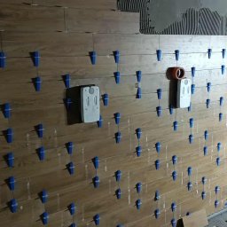 Marcin Domagalski. Usługi budowlane DOM-BUD - Układanie płytek imitujących drewno na ścianie z użyciem niebieskich klipsów poziomujących. Widoczne puszki elektryczne i fragment surowej ściany.